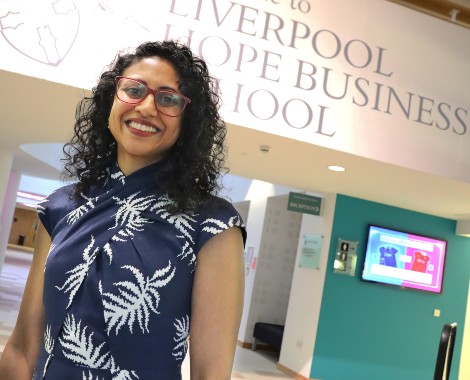A woman wearing glasses and a blue and white dress stands in front of a wall with a Liverpool Hope Business School sign.
